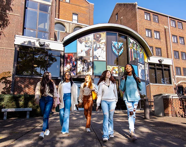A group of five diverse young women walking together in front of a college building with large windows and a display of photographs.